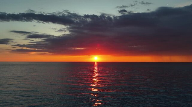 Aerial drone footage pulling backward and upward over a calm sea during sunset. The glowing sun reflects across the water while dramatic clouds and distant rainfall create a striking evening seascape.
