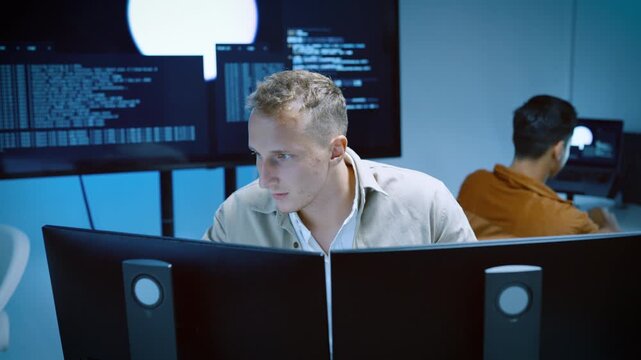 Focused male software engineer analyzing code on multiple monitors in a modern high tech office environment with colleagues working in the background