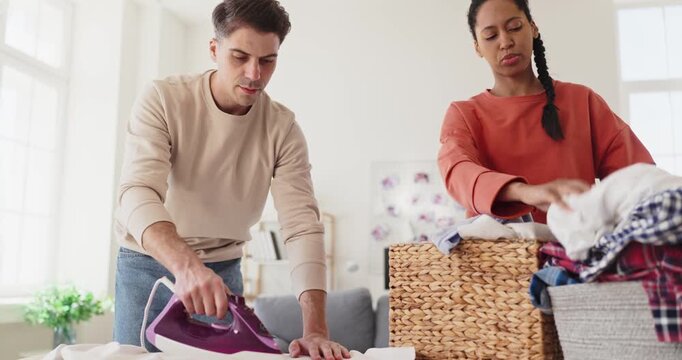 Couple ironing laundry at home, sharing chores. A man uses an iron while a woman sorts clothes into a basket, showing teamwork and efficient housekeeping in a cozy apartment.