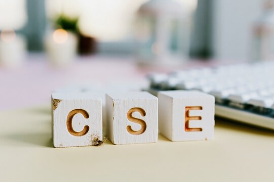 Three white wooden letter blocks spelling CSE on a light desk surface with a keyboard for computer science education, tech industry branding and software engineering