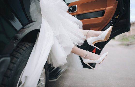 An elegant bride in a wedding dress steps out of a luxury car. A close-up of her legs in white high-heeled shoes is visible.