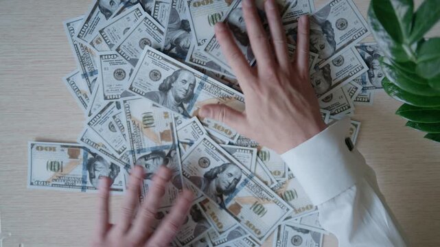Male hands in a white shirt stroking a pile of one-hundred-dollar bills on a wooden table