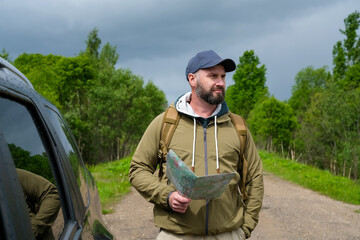 Fototapeta premium Bearded male driver studies paper map beside crossover vehicle on rural road. Concept of road trip planning and outdoor navigation.