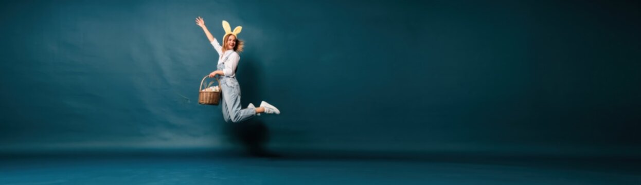 Young woman wearing bunny ears jumping with an easter basket against teal studio background for seasonal holiday marketing, spring advertising and celebratory social media