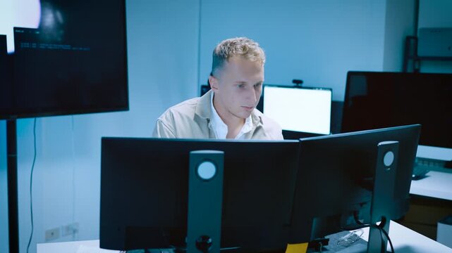 Focused young male IT professional or programmer working with multiple monitors in a high tech office laboratory or server room setting taking notes