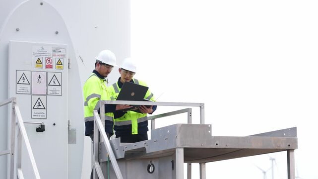 Two Asian industrial workers standing under huge windmills generator field station outdoor. Focused engineering professionals evaluating renewable clean energy power layout.