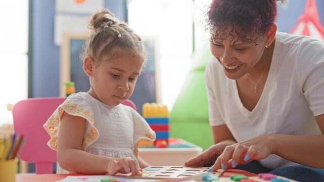 Woman guides little girl fitting wooden puzzle piece at a preschool table inside a building; nurturing.