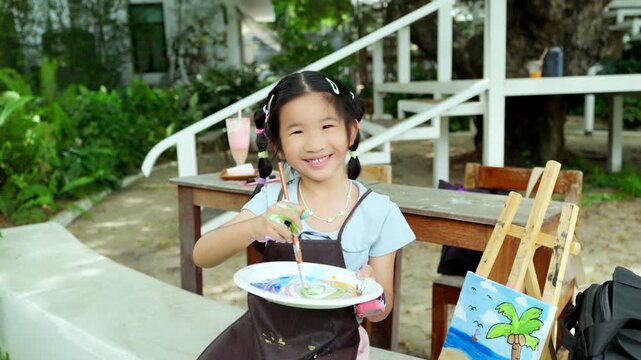 Cute happy Asian little girl showing her colorful painted hand, smiling child enjoying creative art workshop and beach canvas painting activity in the park