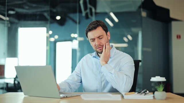 Close-up of modern businessman or IT specialist touching jaw and feeling acute tooth pain or sensitive enamel at computer desk. Overworked corporate manager suffering from dental health problems