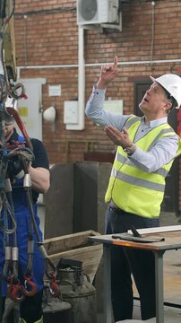 Factory supervisor gives instructions to worker during overhead crane lifting operation in industrial workshop. Teamwork, workplace safety and manufacturing process concept.