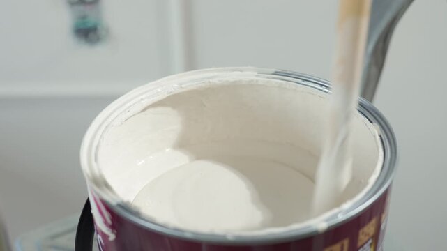 Stirring white paint in an open paint can with a wooden stick. Concept of painting preparation renovation and DIY repair.