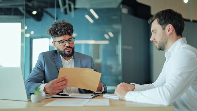 Middle Eastern candidate in suit sitting at desk, showing or giving reference letter in craft envelope to Caucasian employer. Diverse men during hiring process or performance review in modern office.