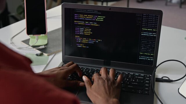 Over the shoulder shot of unknown African American young adult programmer typing code on laptop computer in office. Video for software development marketing, IT education, and tech startup promotions