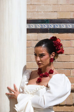 A beautiful woman dancing flamenco, leaning against a white marble column in Seville, Spain. She is wearing a traditional white dress with ruffles and has red flowers in her dark hair.