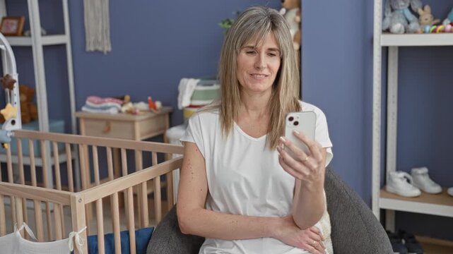 Woman middle age blonde caucasian holding smartphone by wooden cradle, smiling and looking down while seated in an armchair with nursery shelves, toys and dresser in a building; calm bonding.