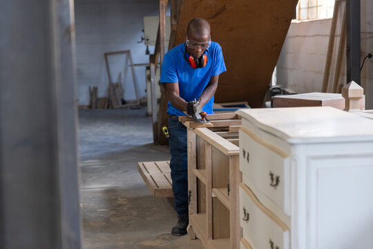 Craftsman planing cabinet frame in wood shop, with lumber stacks and white dresser near window