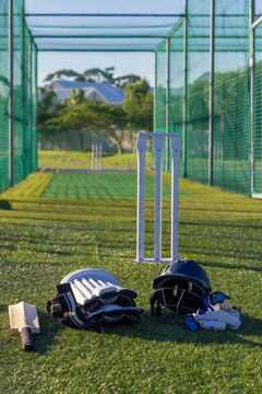 Cricket batting kit is lying on green turf inside practice net, sunlight is casting long shadows