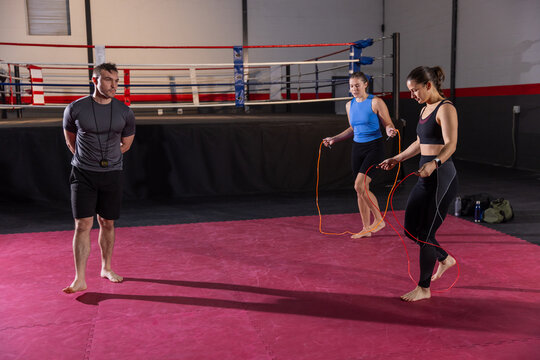 Male coach with whistle timer watching two women trainees skipping ropes barefoot on mat near ring