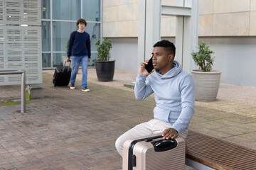 Diverse male travelers sitting on bench at plaza speaking on smartphone beside beige suitcase © wavebreak3