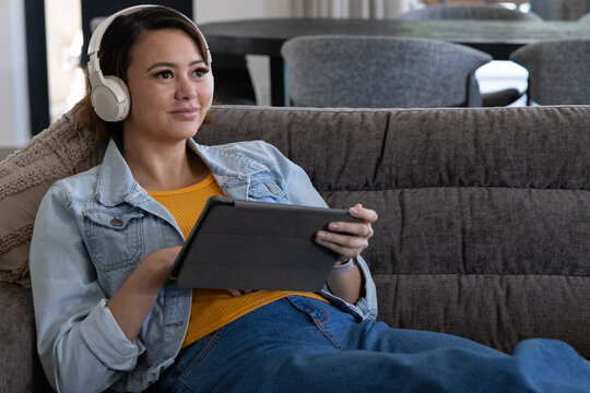 Adult African American woman reclining on gray tufted sofa wearing headphones using tablet