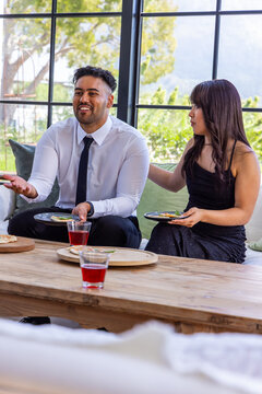 Diverse couple sitting on sofa in lounge wearing formal attire holding plates near coffee table
