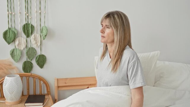 Woman sitting up in bed with eyes closed beside a bedside table and lamp in a bright bedroom; quiet contemplation.