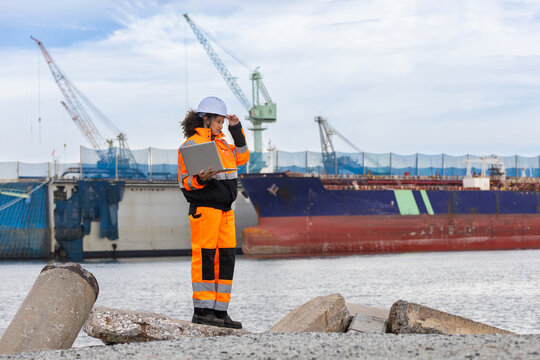 Female engineer with laptop at shipyard terminal, Logistics coordinator monitoring shipping vessel operations at a commercial harbor