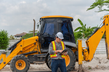 Construction engineer wearing safety gear using tablet beside yellow excavator at worksite highlighting project management equipment inspection site operations © JP.Studio