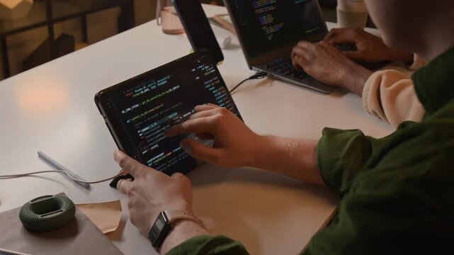 Over the shoulder shot of young programmer scrolling through software code on tablet screen while colleague working on laptop. Ideal for marketing materials promoting mobile app development services