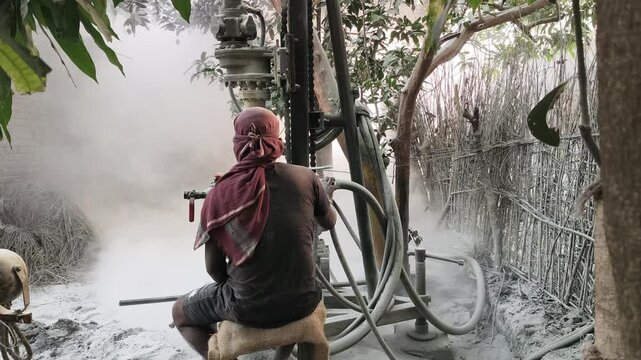 A worker with a red cloth head covering operates a borewell drilling rig outdoors. A thick cloud of white dust fills the air, covering the machinery and a nearby stick fence. 