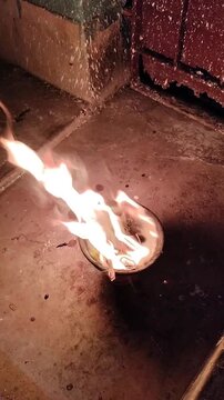 high-angle, close-up shot of a small bowl containing a bright, dancing fire at night. The flames flicker and sway, casting a warm, glowing light against the dark, textured ground. This atmospheric foo