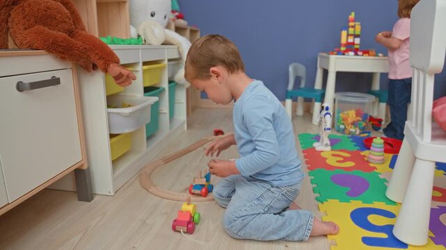 Boy kneeling on floor builds wooden train track and pushes a toy car beside foam puzzle mat and storage shelves with stuffed animals in building; playful childhood joy.
