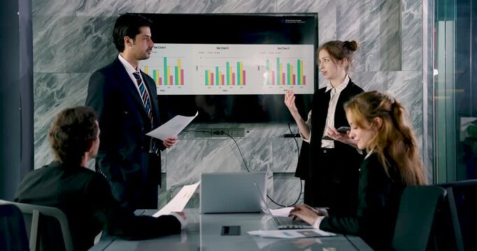 Businessman holding document explaining chart while businesswoman prepares notes during team presentation, group of professionals seated at table focusing on data-driven discussion