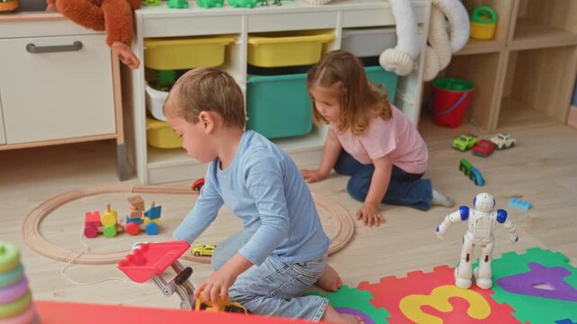 Boy and girl building block tower and steering toy truck on floor in a playroom with shelves of stuffed animals; playful cooperation.