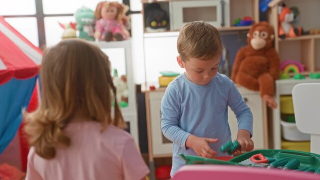 Boy holding a green plastic toy mirror and turning its handle while a little girl watches from behind a table in a brightly lit playroom building; curiosity sharing.