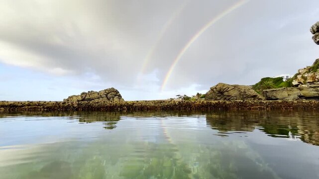 Clear water in tidal pool with a double rainbow in sky on a cloudy morning.