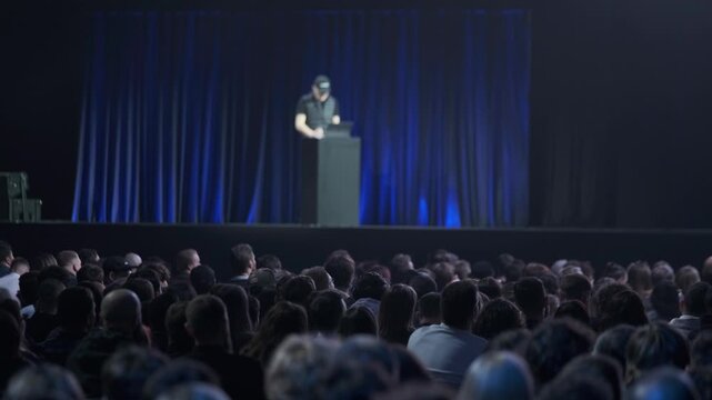 Large audience watches speaker on stage during conference presentation, blue curtain backdrop and dim lighting.