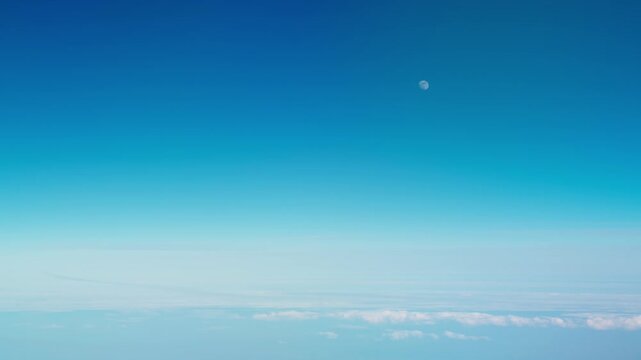 Passenger plane window seat view of daytime moon and blue sky over clouds inflight, handheld
