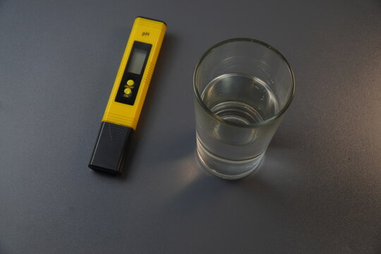 Yellow digital pH meter tester pen next to a clear glass of fresh water on a grey background. Concept of water quality testing, acidity measurement, chemical laboratory, and purification control.