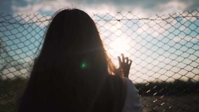 Woman pressing hand on chainlink fence at cloudy sunset outdoors. Girl touches fence barrier with hand. Silhouette shows woman at chainlink. Girl pressing wire fence under cloudy sky view.