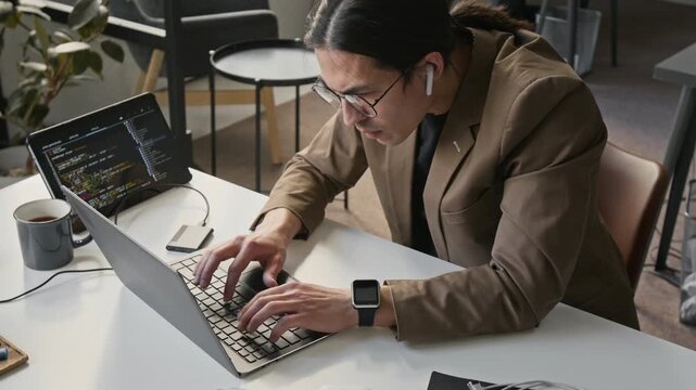 Asian young programmer wearing glasses coding on laptop with dual screen setup in office. Footage for tech startup marketing, software development, or IT education