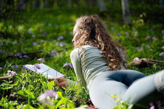 Beautiful female reader lying on flower meadow in forest with book, mindfulness, education and peaceful outdoor recreation, back view.
