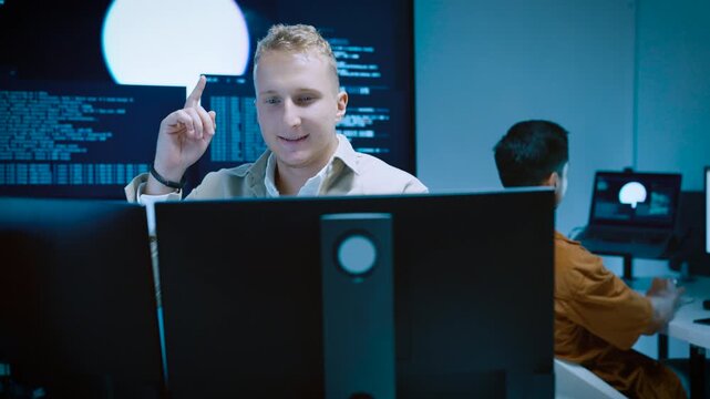 Young male programmer raising finger with creative idea while coding and analyzing data on computer monitors in tech office
