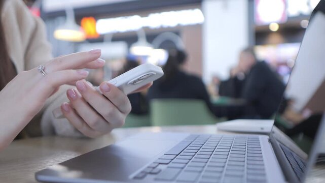 Person uses smartphone beside laptop in cafe. Fingers scroll actively on phone screen. Background shows blurred cafe patrons. Ideal for tech, communication, or multitasking themes