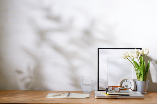 Minimal wooden desk with white books, tulip flowers, eyeglasses, notebook, headphones, and blank frame against a bright wall with soft shadows.