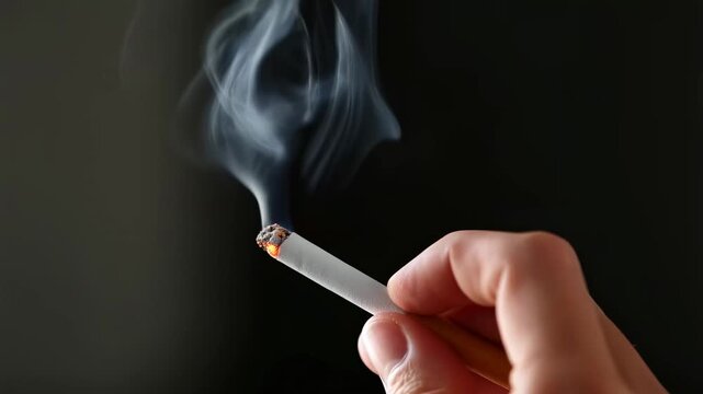 Close-up of a hand holding a burning cigarette with smoke rising against a dark background. Represents smoking culture and its health implications