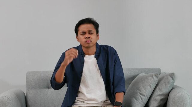 A young man sits on the sofa in the living room at home, with a confused expression while scratching and shaking his head, raising his hands and shoulders hesitantly, looking at the camera.