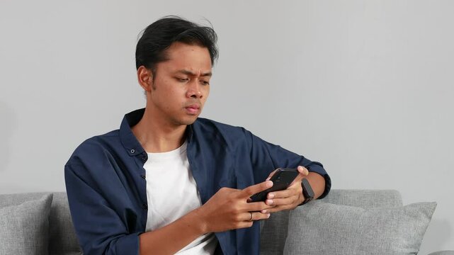 A young man uses a smartphone while sitting on a sofa in the living room, surfing the internet, using social networks, shaking his head with an expression of shock at the bad news and disbelief.