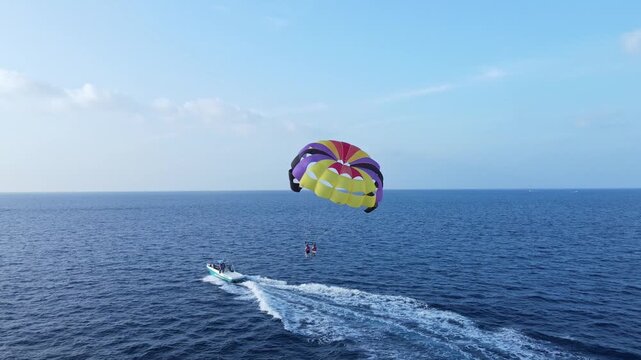 Drone view of two tourists parasailing above deep blue ocean waters while being towed by a speedboat. Tropical adventure, water sports, and vacation travel concept.