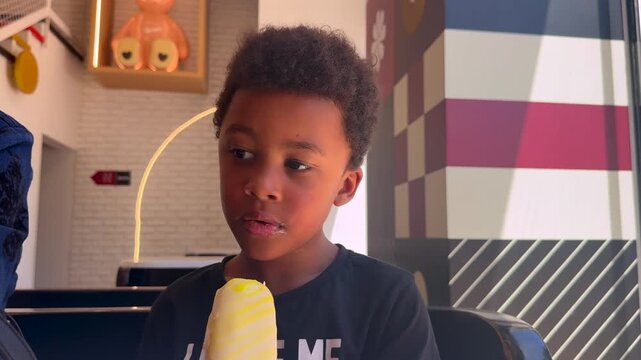 6 year-old biracial black child delighted eating a vanilla sa ice cream indoor. he is wearing curly afro style hair and very expressive black eyes.